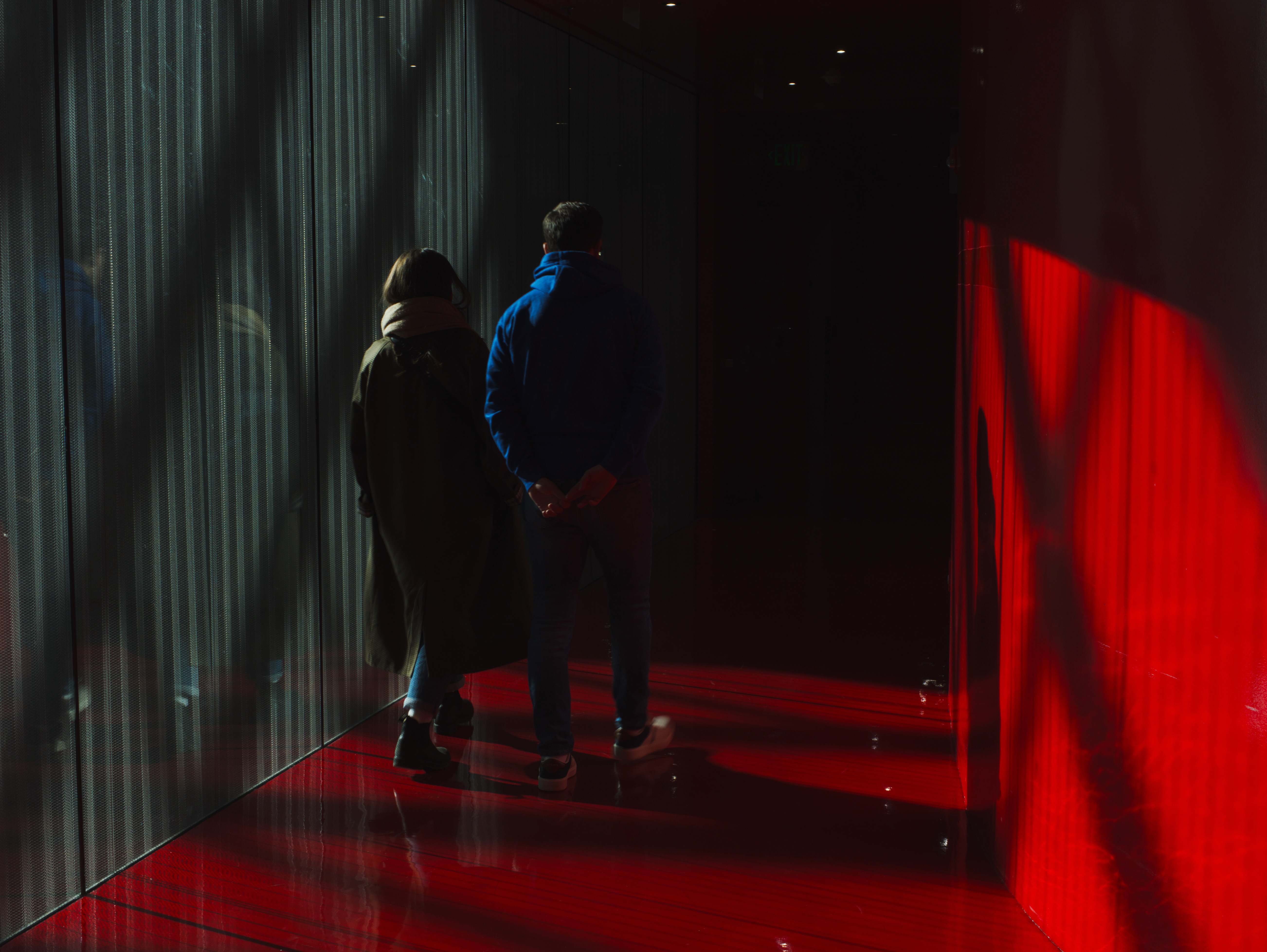Two people stand in a dimly lit corridor with green vertical panels on the left and glowing red light on the right, viewed from behind.