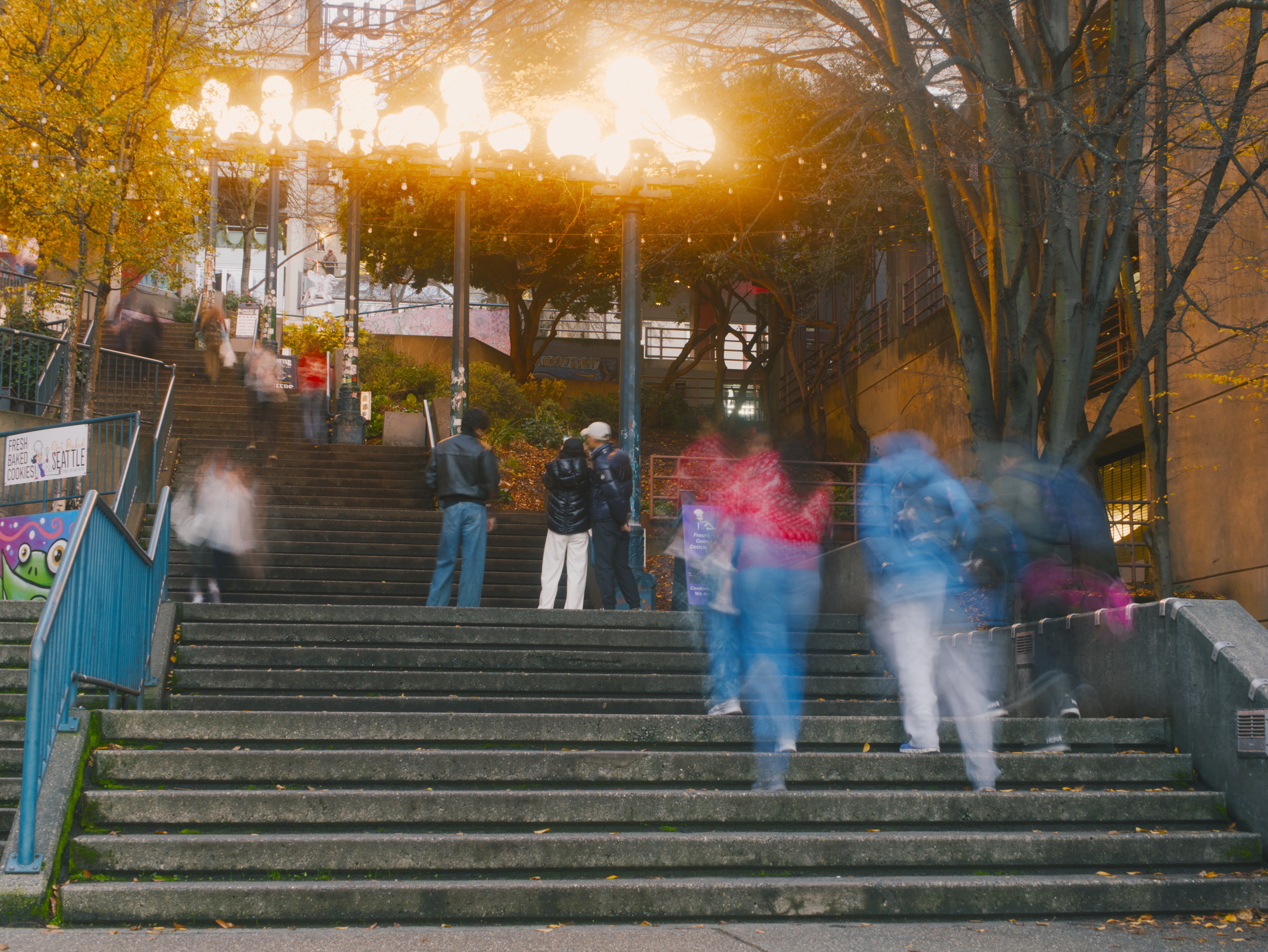 People walk up and down a long outdoor staircase at dusk, with some figures motion-blurred while bright street lights glow above in the autumn trees.