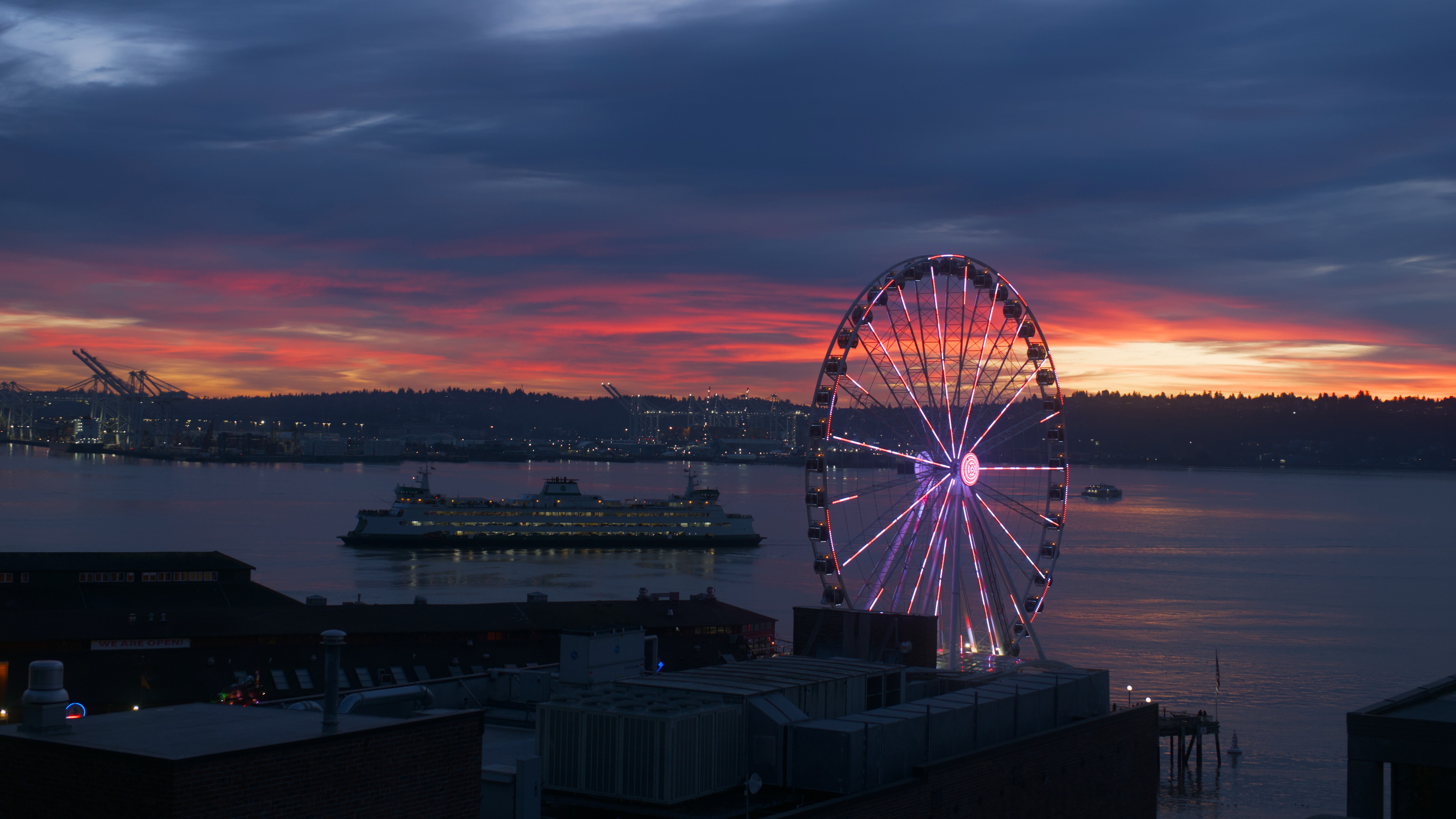 A brightly illuminated Ferris wheel on a waterfront pier glows with pink and purple lights against a dramatic sunset sky as a ferry passes by in the water.